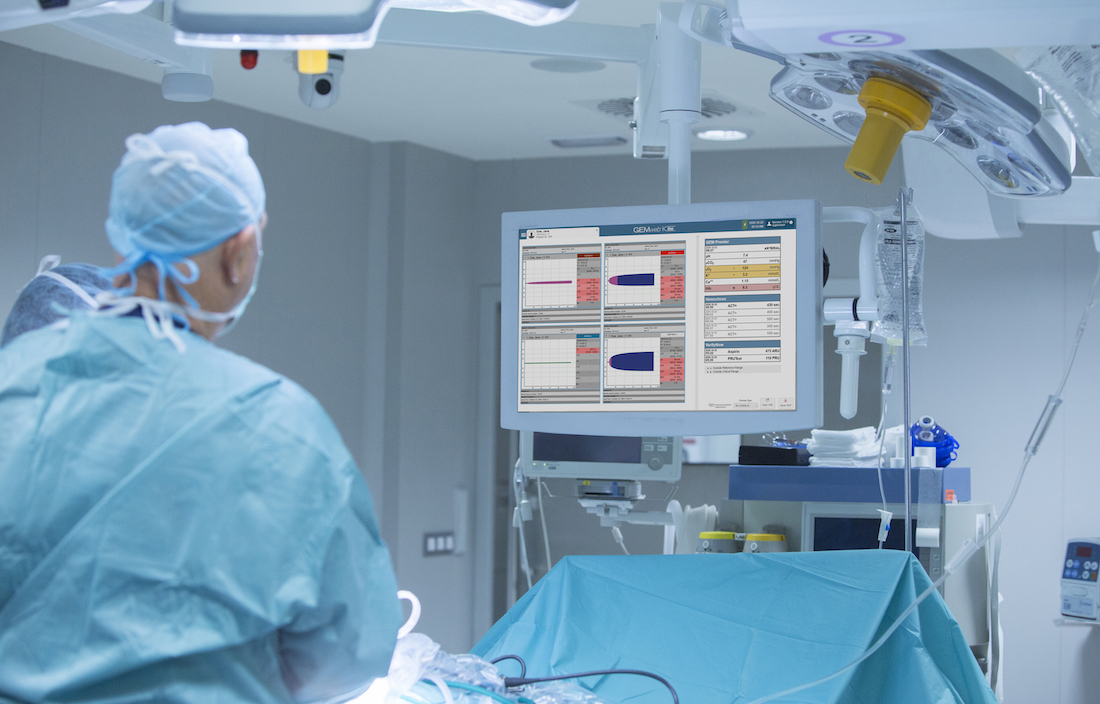 Photo shows a clinician in a CVOR looking at a monitor above the patient, using ROTEM live it shows real-time data about the patient's blood clotting ability.